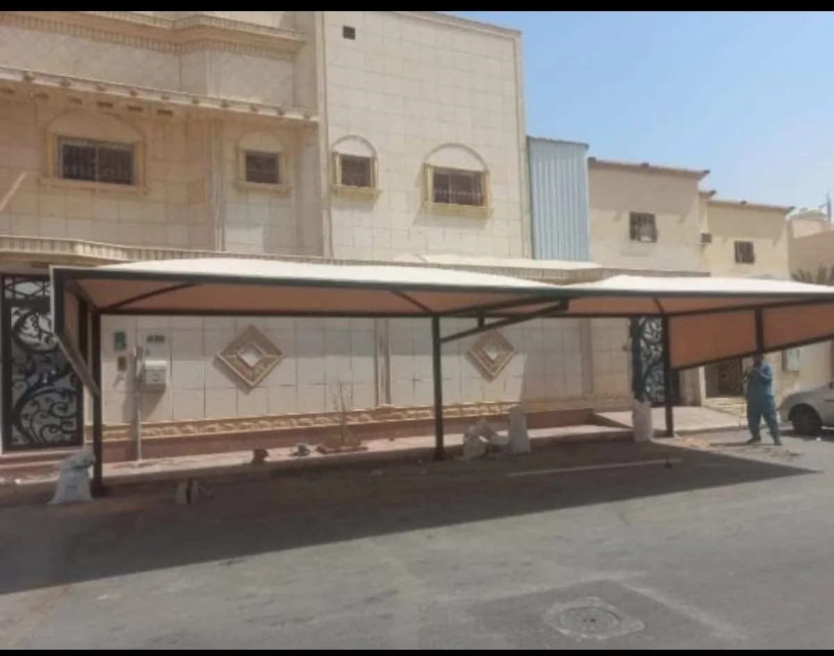 مظلة سيارات بهيكل معدني وقماش فاتح تمتد أمام واجهة منزل حجري