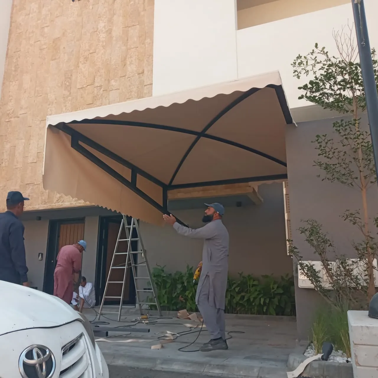 ثلاثة عمال يركّبون مظلة سيارات مقوسة أمام منزل باستخدام سلم معدني